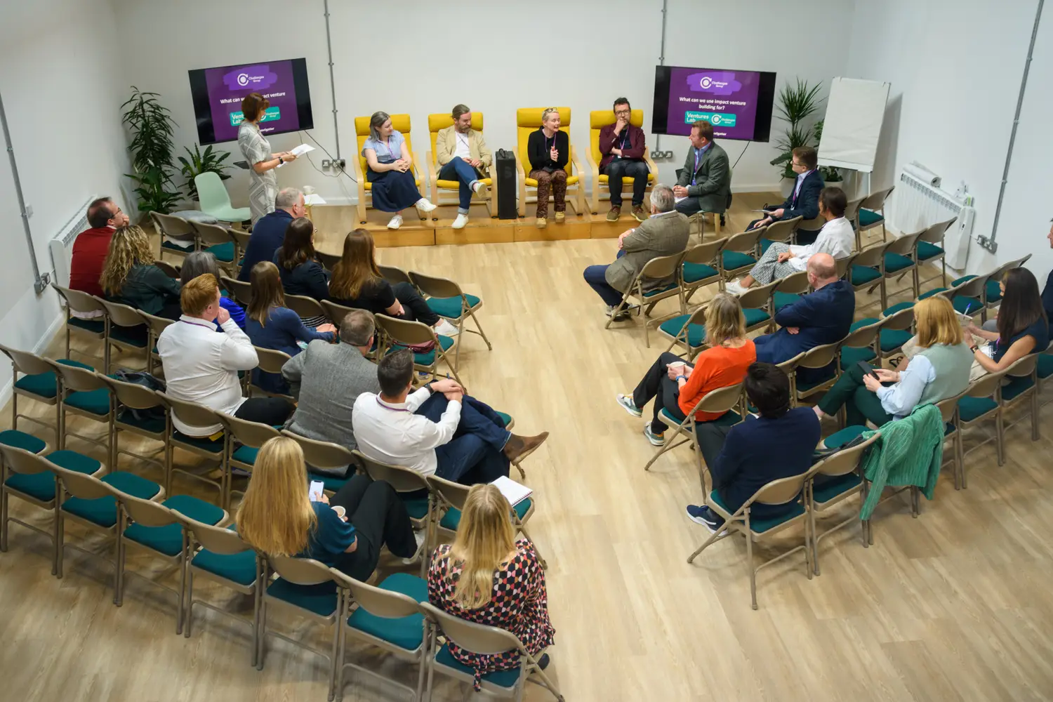 Four presenters sit on yellow armchairs next to two presentation screens. They are facing an audience of people sat on pale brown fold-up chairs.