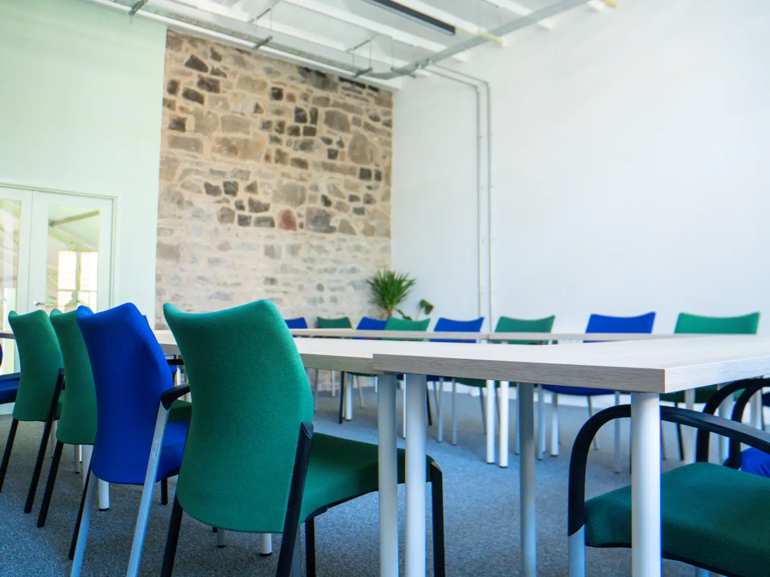A room with a large rectangular shape made up of tables with green and blue chairs. Most of the walls are painted in light colours except one in which you can see the traditional stone wall.