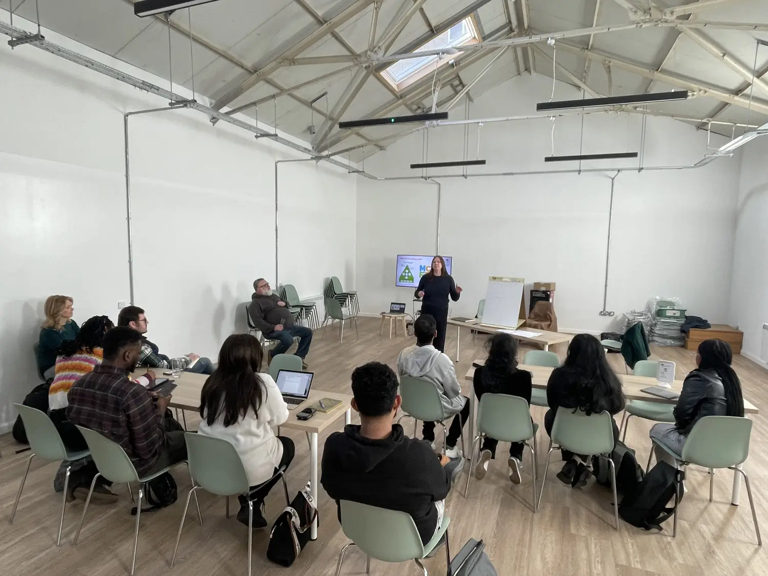 A group of people are seated at tables and chairs in a high roofed room with white walls. A woman addresses them from the back of the room using a presentation screen.