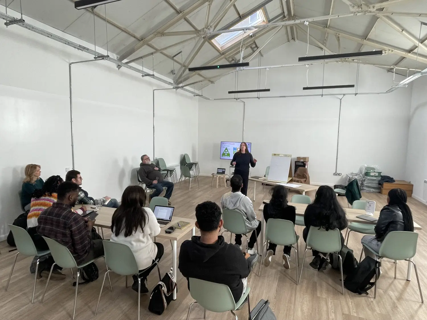 A group of people are seated at tables and chairs in a high roofed room with white walls. A woman addresses them from the back of the room using a presentation screen.