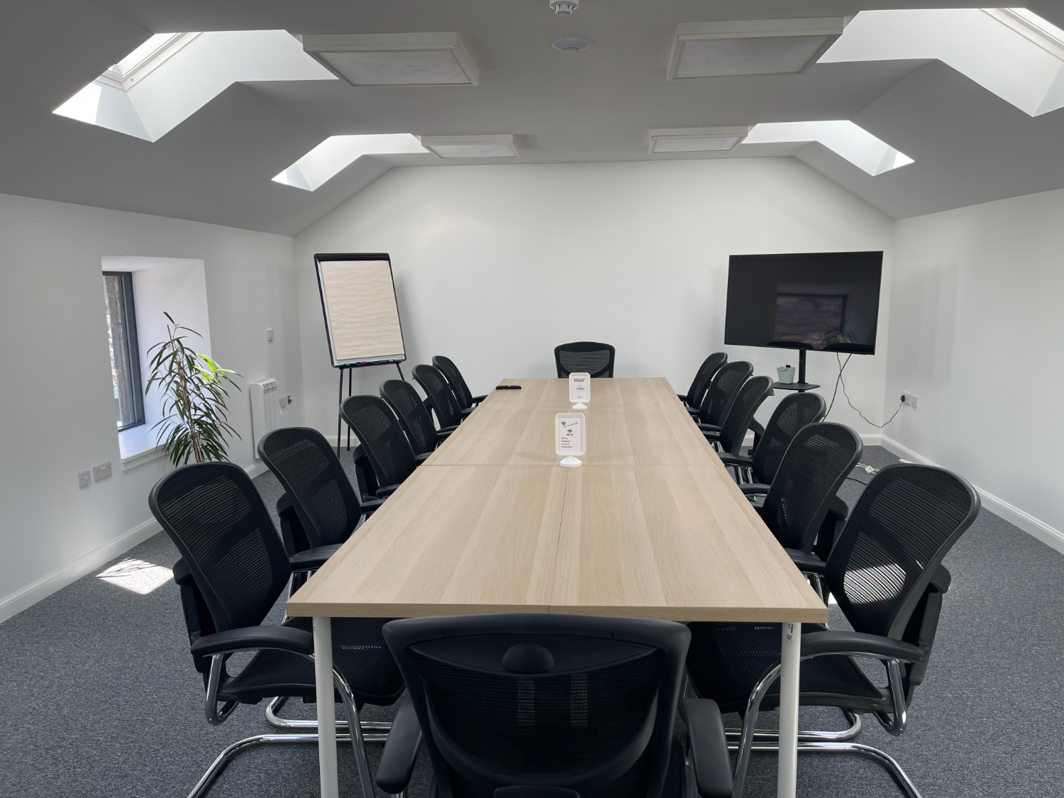 Photo of the "Proving Room" with a large board table, 14 black chairs, grey carpet and a white walls. A screen on a stand and a whiteboard can be seen at the back of the room.
