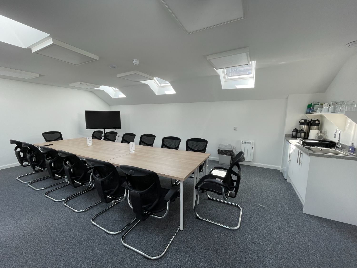 Photo of the "Proving Room" with a large board table, 14 black chairs, grey carpet and a white walls. A small kitchen area can be seen on the right of the photo.