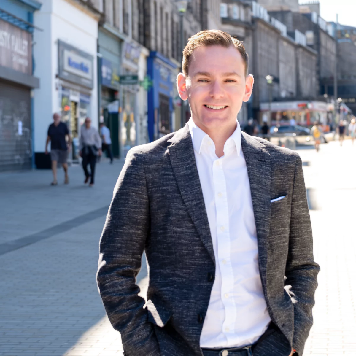 Man stood on a street in Edinburgh with brown hair, fair skin, a blazer style jacket and a white shirt.