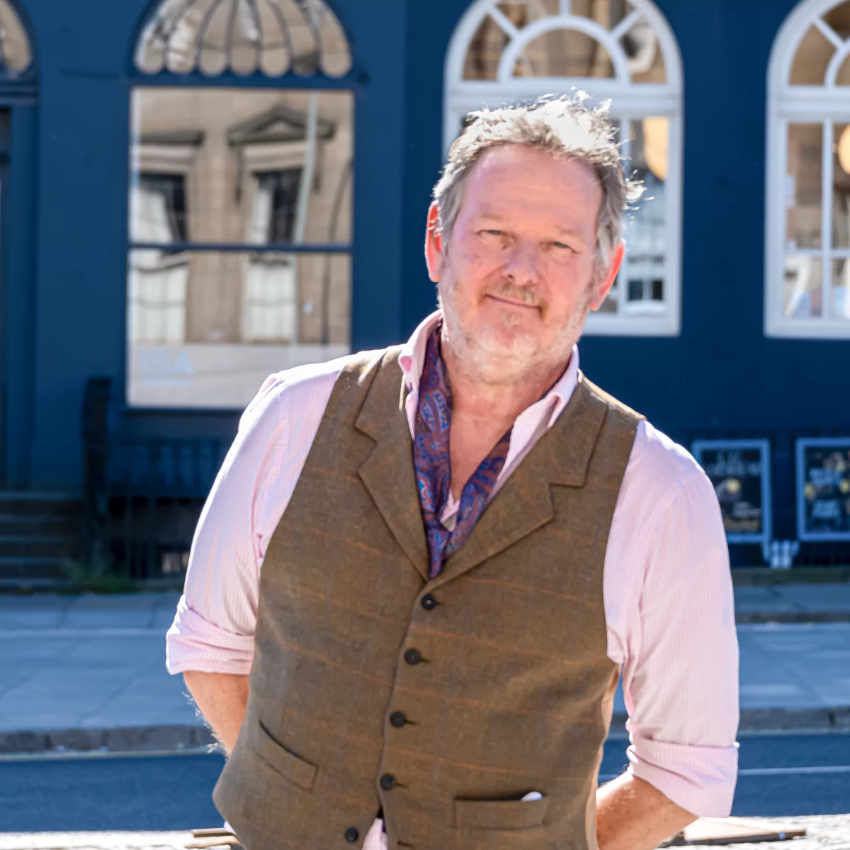 Male with grey hair wearing a brown waistcoat, pink shirt and purple scarf standing in front of a blue building in Edinburgh