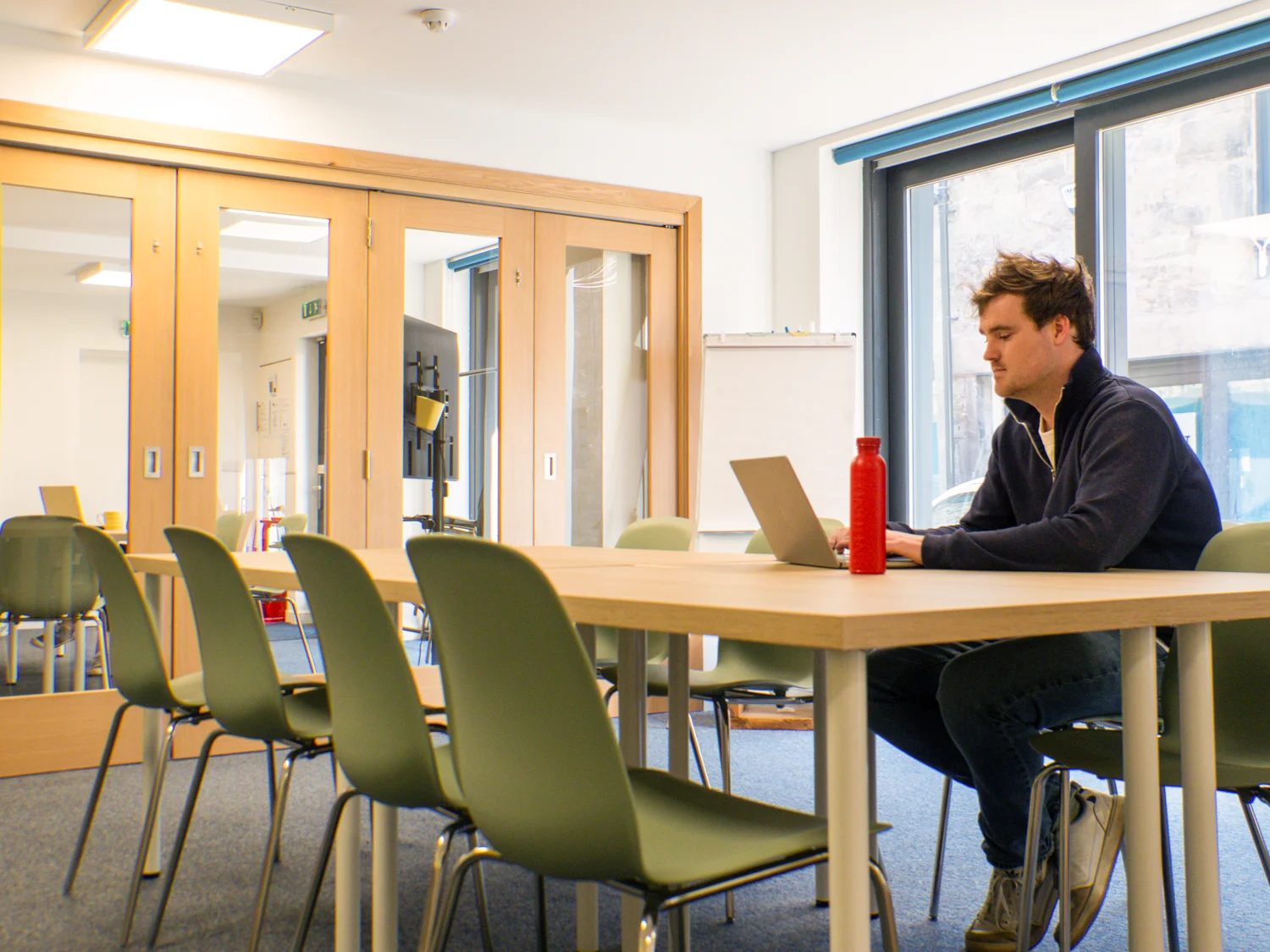 Photo of a meeting room showing a table and chairs with a large window in the background. A white male is sitting at the table using a laptop.